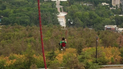 A young man in gear descends on a zip line over a park with the urban backdrop Stock Footage 248529913