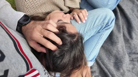 A young man gently caresses his girlfriend’s head as she rests on his lap Видео 332706556