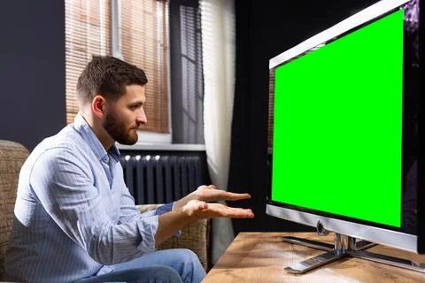 A young man gesticulating in front of TV at home, confused, does not understand Stock Photos