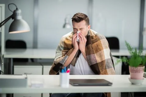 Young man gets sick at work and blows his nose with a napkin at the office desk Stock Photos