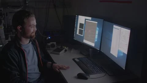 Young man gives comment, looking at the camera, sitting at computer in the dark. Stock Photos
