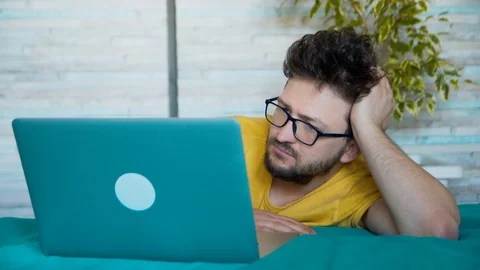 A young man in glasses and with a beard looks lazily at the laptop screen. The Stock Footage 128456387