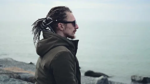 Young man in glasses and dreadlocks hairstyle hair looks into endless sea Stock Footage 236485170