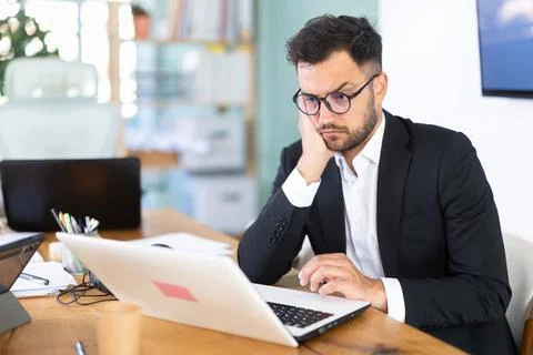 Young man in glasses sits at a table and looks at a laptop screen Stock Photos