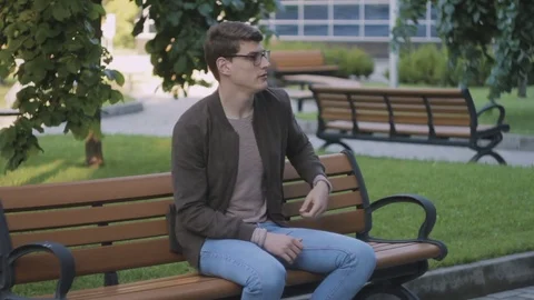 A young man in glasses is sitting on a bench near the building counting bills. Stock Footage 79667688