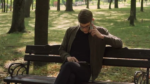 A young man with glasses sitting on a bench in the park works in an application Stock Footage 79693653