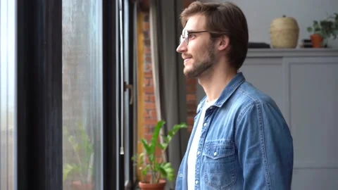 Young man in glasses standing at window at home looking away Stock Footage 142876982