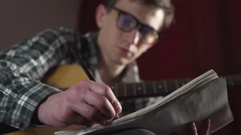 A young man with glasses writes the text of a song with an acoustic guitar in a Stock Footage 236689457