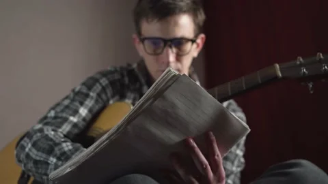 A young man with glasses writes the text of a song with an acoustic guitar in a Stock Footage 237017419