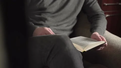 Young man in gray sweater reading a book sitting in an armchair at home. Stock Footage 235530123