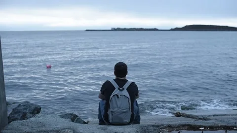 Young man with grey backpack is sitting on the pier and watching the slow motion Stock Footage 233193187