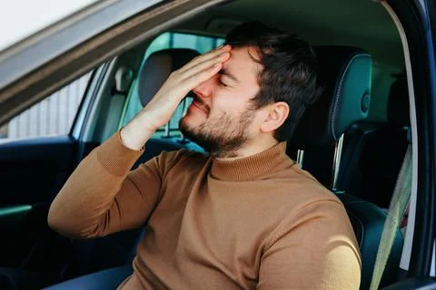 A young man guy sits in a car holding his hand on his head which hurts. Foto stock