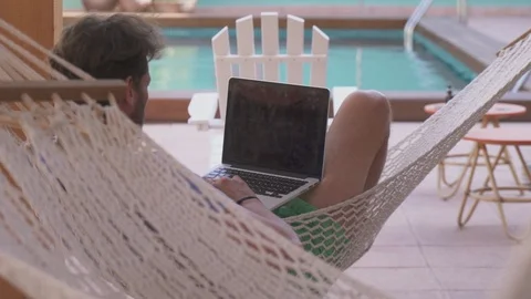 A young man in a hammock by the pool working on a laptop Stock Footage 100396795