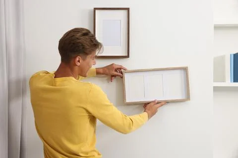 Young man hanging picture frames on white wall indoors, back view Stock Photos