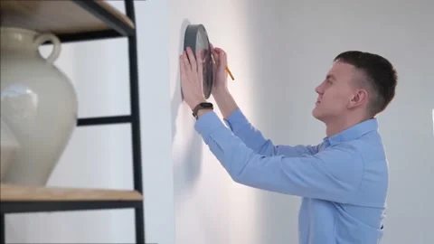 Young man hangs a clock on an empty white wall in his first new home Stock Footage 168184759