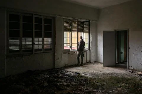 Young man with hat and jacket watching out of an old abandoned building windowsi Stock Photos