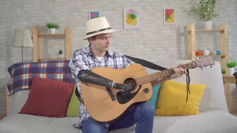 Young man in a hat with a bionic prosthetic arm plays an acoustic guitar sitting Stock Footage 119275612
