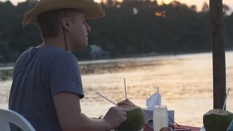 Young man in a hat drinking a coconut in a beach cafe. outdoors. Coconut water Stock Footage 200071116