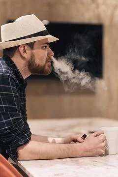 Young man in hat drinking coffee and smoking an electronic cigarette during.. Stock Photos