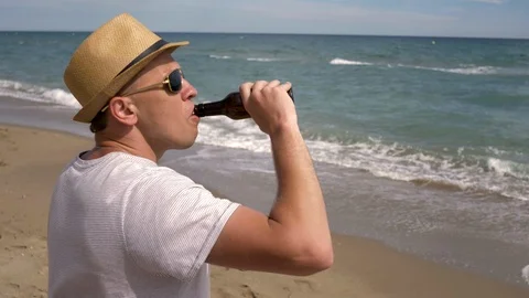 A young man in a hat drinks beer from a brown bottle while standing by the sea Stock Footage 120871407