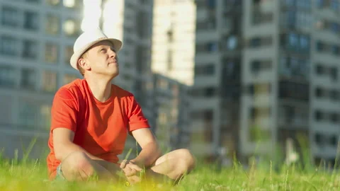 Young man in a hat resting sitting on the grass in a city park, the camera Stock Footage 143840218