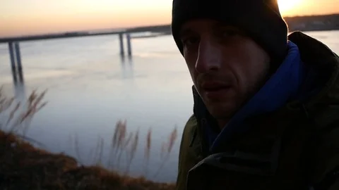A young man in a hat on the river at a bridge with a sunset, does selfie, looks Stock Footage 82297043