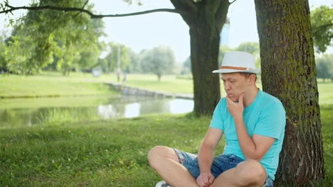 Young man in a hat sits on the shore of the lake under a tree, copy space Stock Footage 138459630