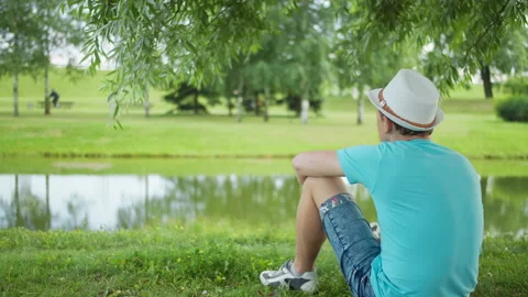 Young man in a hat sits on the shore in a city park, back view Stock Footage 148555983