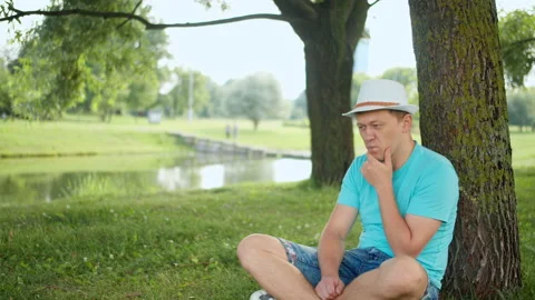 Young man in hat sits thoughtfully under a tree in the park, tracking the camera Stock Footage 138632218