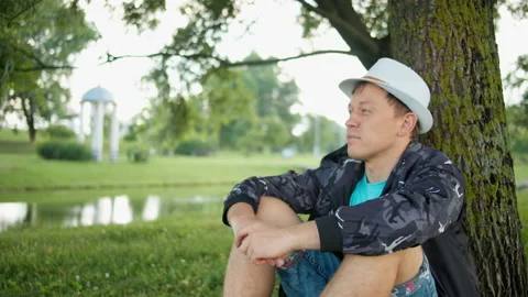 Young man in a hat sits under a tree in the park, camera movement, close-up Stock Footage 138504866