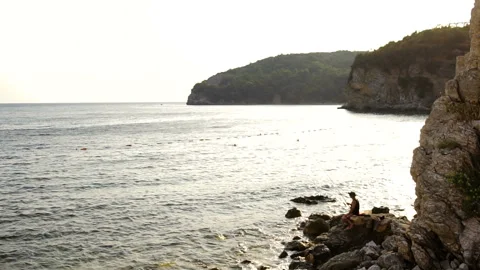 Young man in a hat sitting on the rocks and fishing at the sea coast with cliffs Stock Footage 85643373