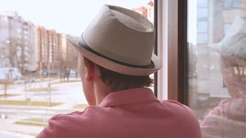 Young man in a hat standing in the window opening looks at the street and drinks Stock Footage 158701566