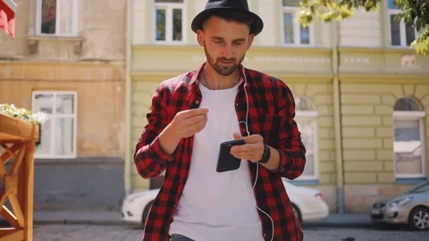 Young man in the hat uses phone listen music headphone walk in city smile feel Stock Footage 92070733