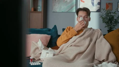Young man having cold is sitting on sofa, watching TV, sneezing into his Stock Footage 153092000