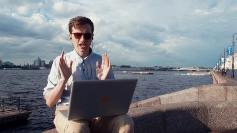 Young man having video call at laptop sitting on bench near seafront. Summer day Stock Footage 77167583