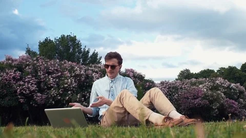 Young man having video call at laptop sitting on green grass at park. Summer day Stock Footage 77168177