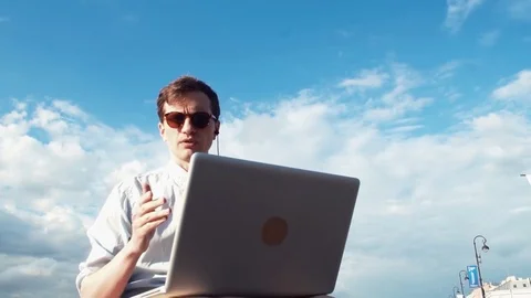 Young man having video chat with laptop sitting on bench outdoors. Summer day Stock Footage 77167638