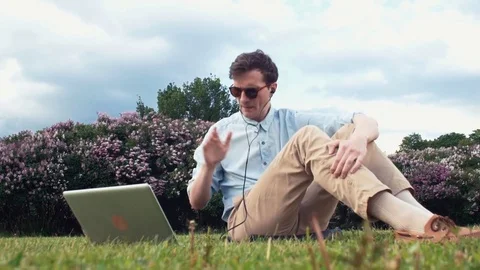Young man having video chat at laptop sitting on green grass at park. Summer day Stock Footage 77168239