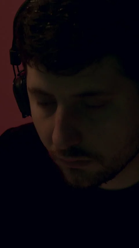 Young man with headphones focused on work in a dimly lit room, illuminated by Stock Footage 315441981