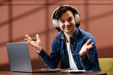 Young man with headphones gestures while sitting at a desk with a laptop, p.. Foto stock