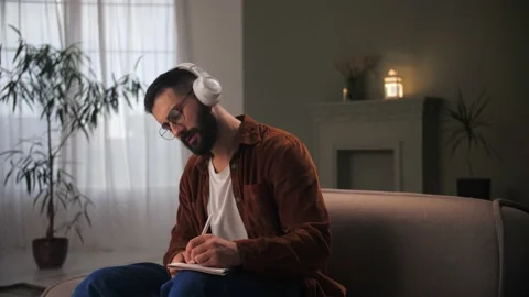 Young man with headphones typing notepad using pen and paper.Bearded guy doing Stock Footage 212315692