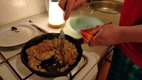A young man in his home kitchen stirs buckwheat in a frying pan Stock Footage 307496489