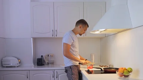 A young man in his kitchen, cut tomatoes and fry it. Man prepares food and Vidéo 114273704