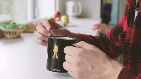 Young man is holding a cup of tea or coffee with his hands and stirring sugar wi 스톡 동영상 223422069