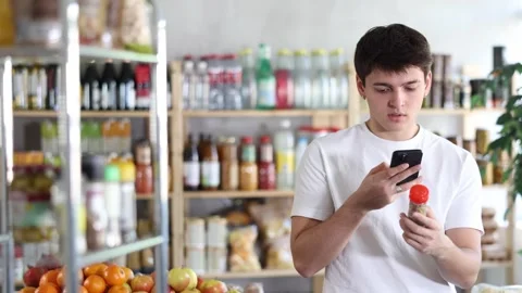 Young man holding jars with spices, using smartphone to scan label on cardamom Stock Footage 341533274