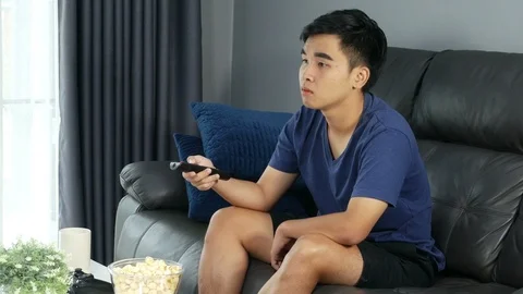 Young man holding remote control and watching TV while sitting on sofa Stock-Footage 93418356