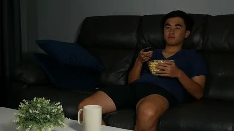 Young man holding remote control and watching TV while sitting on sofa Video stock 93421902