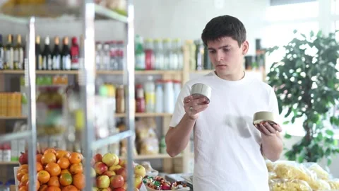 Young man holding two tin cans of preserves in hands, choosing and shop around Stock Footage 313874370