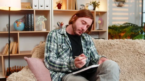 Young man on home sofa thinking hard then writing notes in notebook after good Stock Footage 347051736