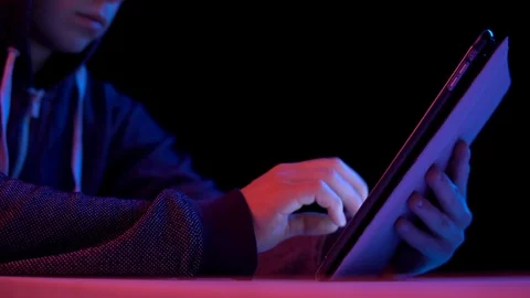 Young man in a hood with a tablet. Hacker makes a hack through the tablet. Blue Stock Footage 125402026